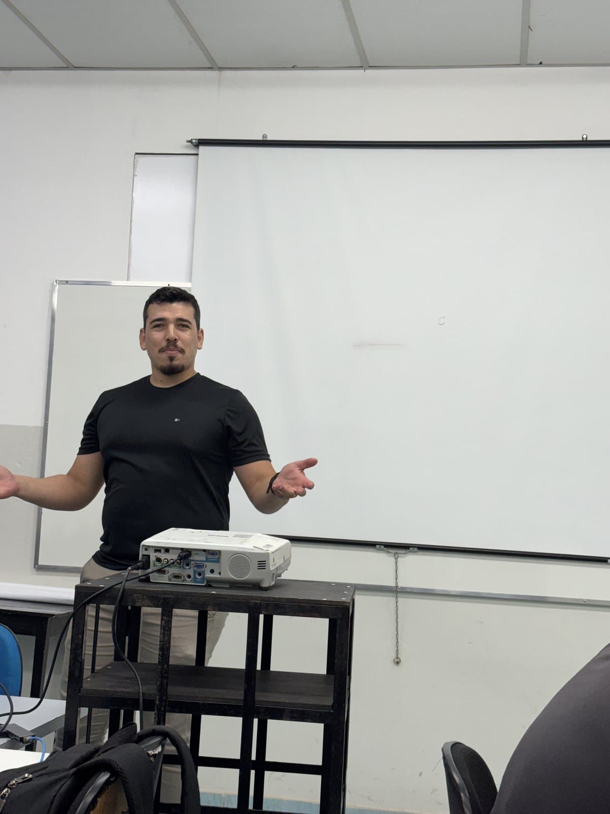 João Camilo em sala de aula, em frente ao quadro, ao lado do projetor